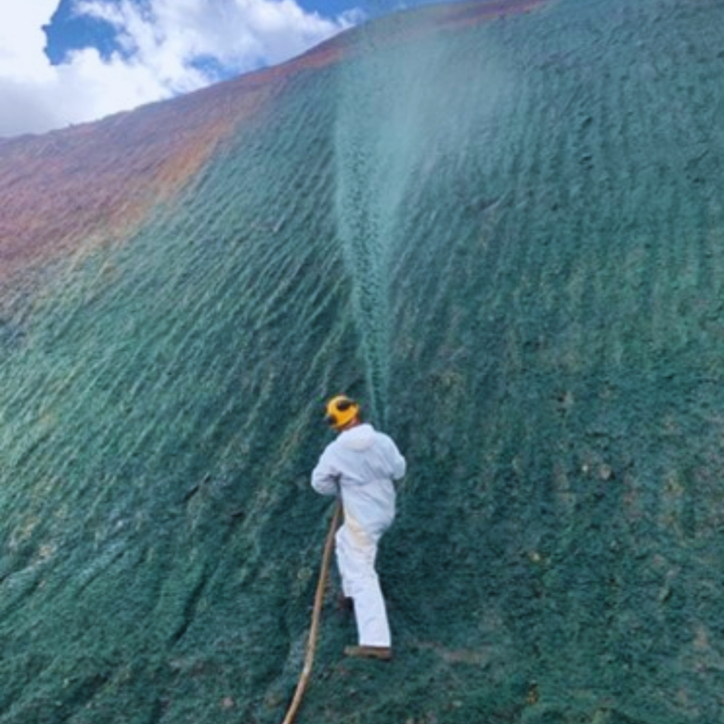Homem com uniforme branco aplica Hidroplantio por jateamento em talude de alta inclinação, durante processo de recuperação de área degradada em mineradora.